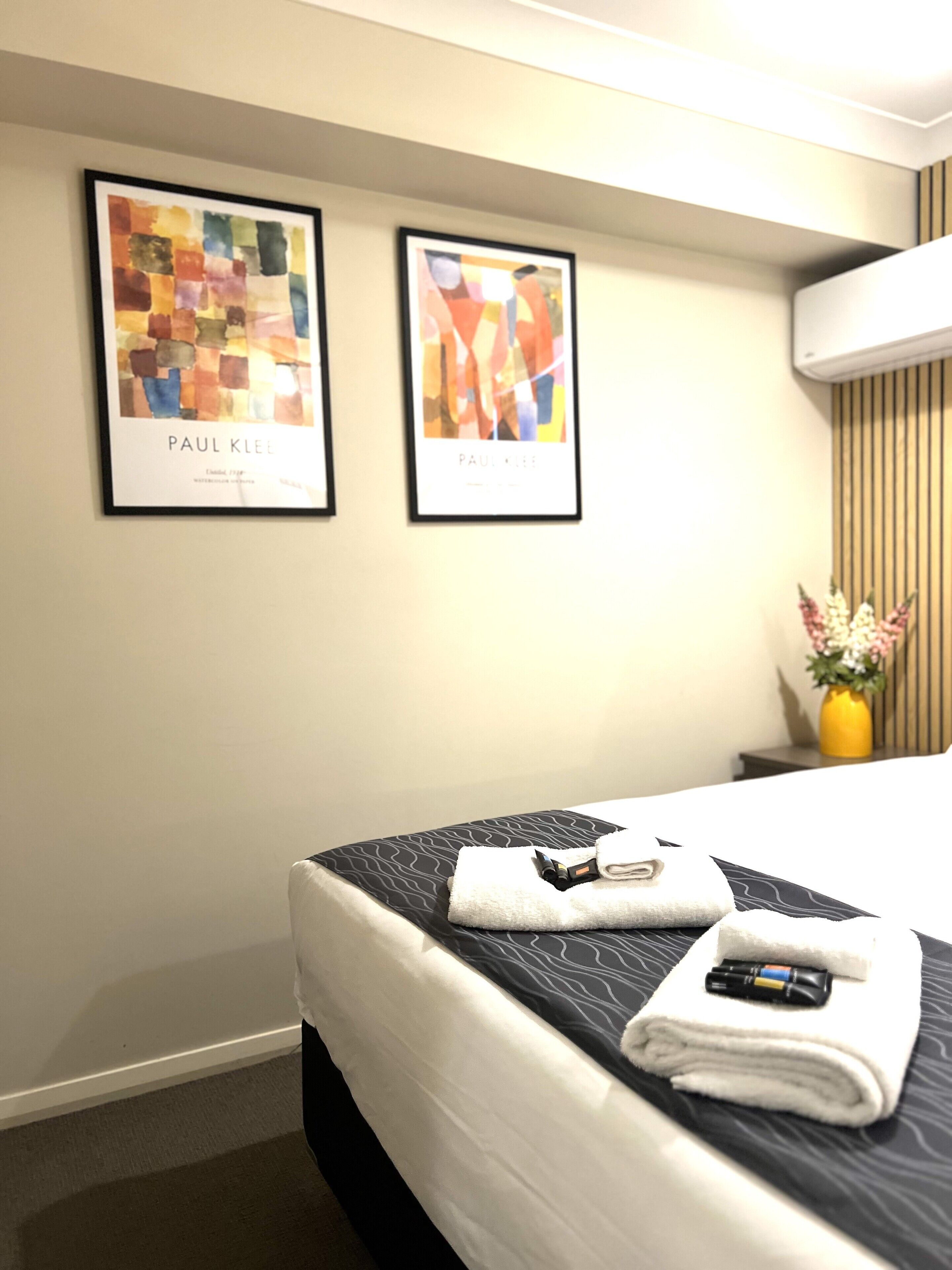 A hotel room with a neatly made bed, two framed artworks on the wall, a bedside table with a vase of flowers, and a wall-mounted air conditioner.