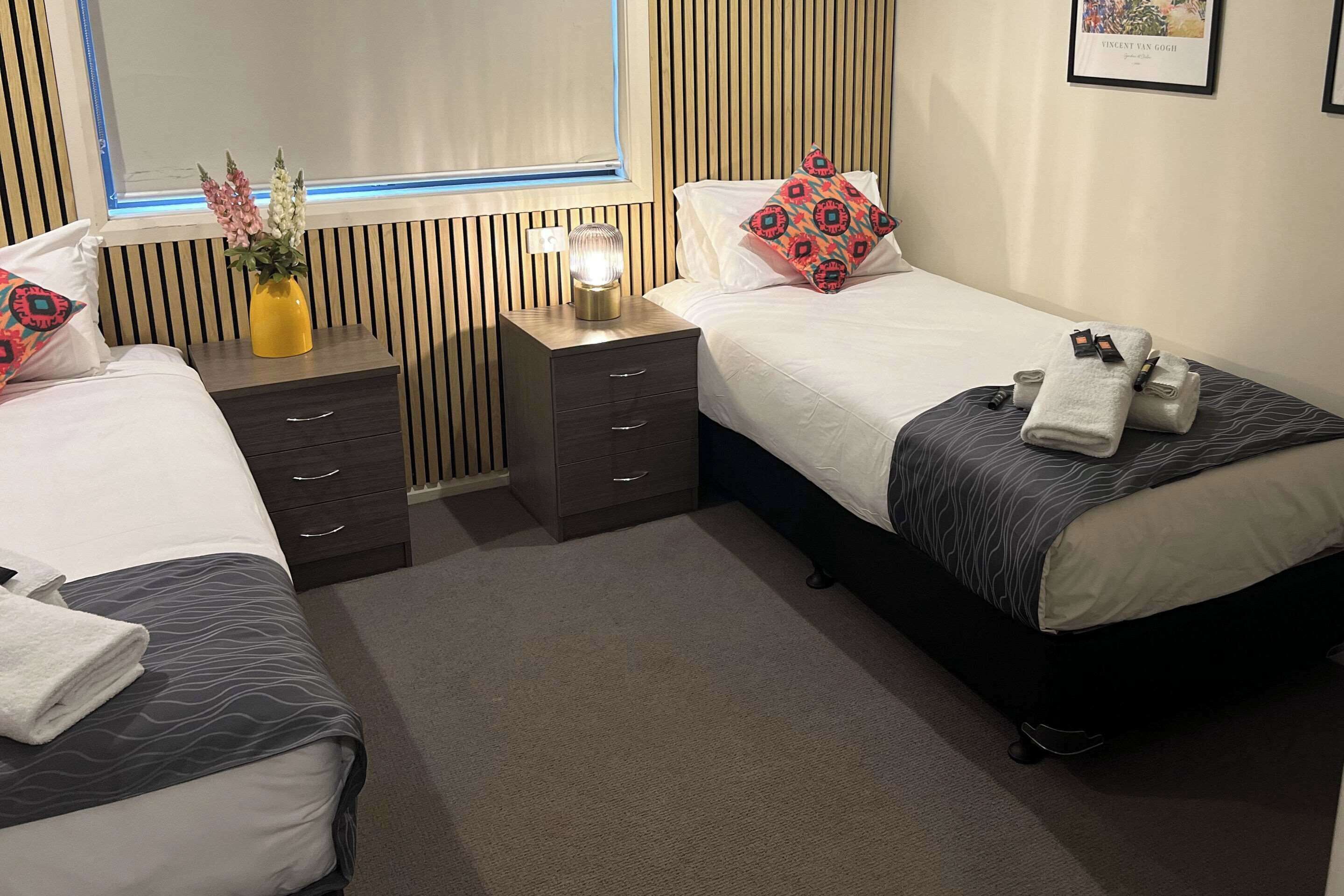 A hotel room with two beds, a nightstand with drawers, a vase with flowers, and a framed picture on the wall.