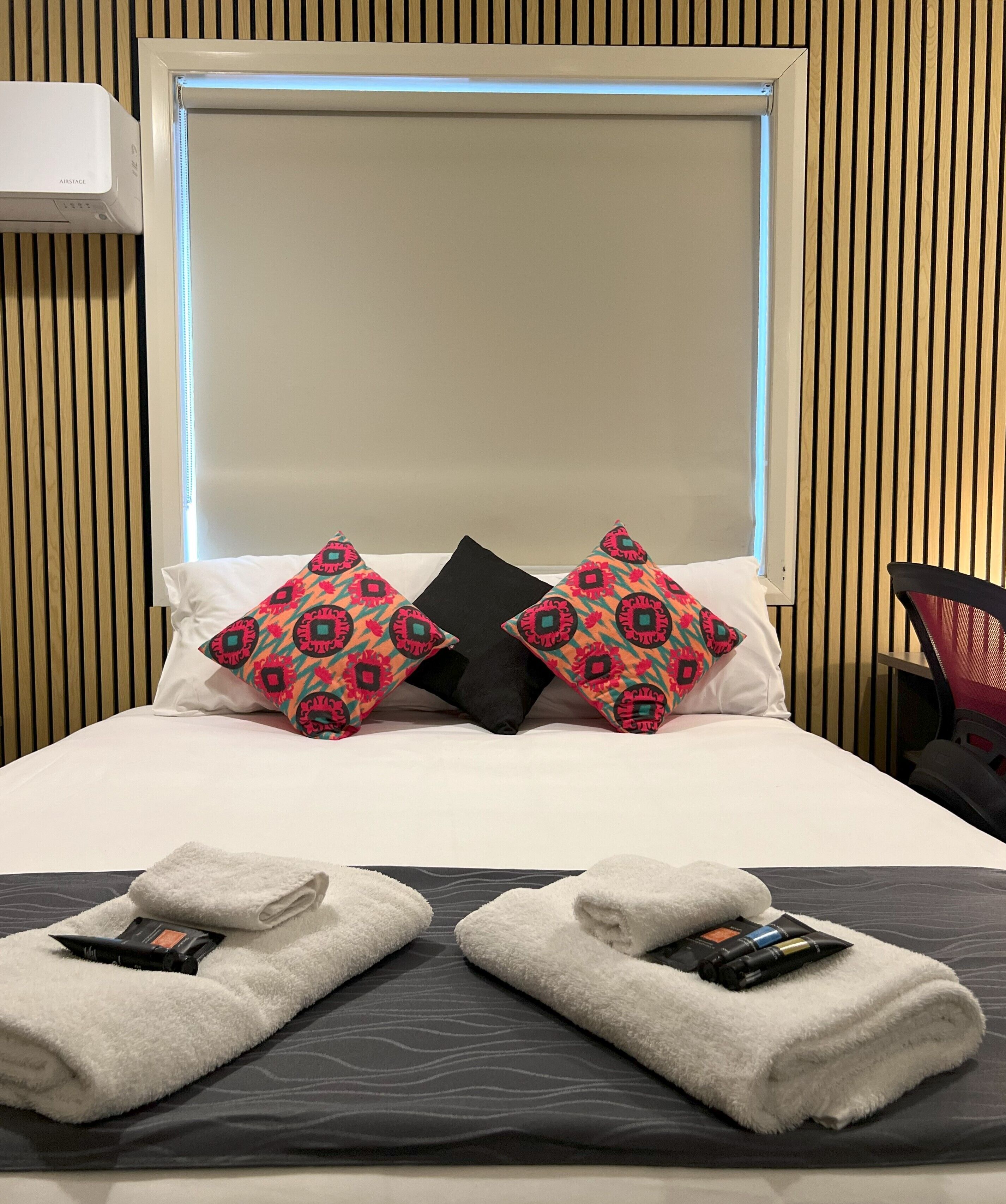 A hotel room with a bed, two decorative pillows, a bedside table with towels and toiletries, and a chair.