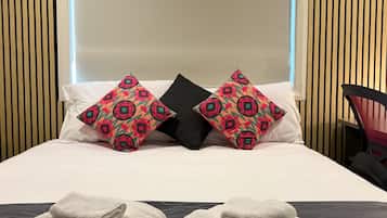 A hotel room with a bed, two decorative pillows, a bedside table with towels and toiletries, and a chair.