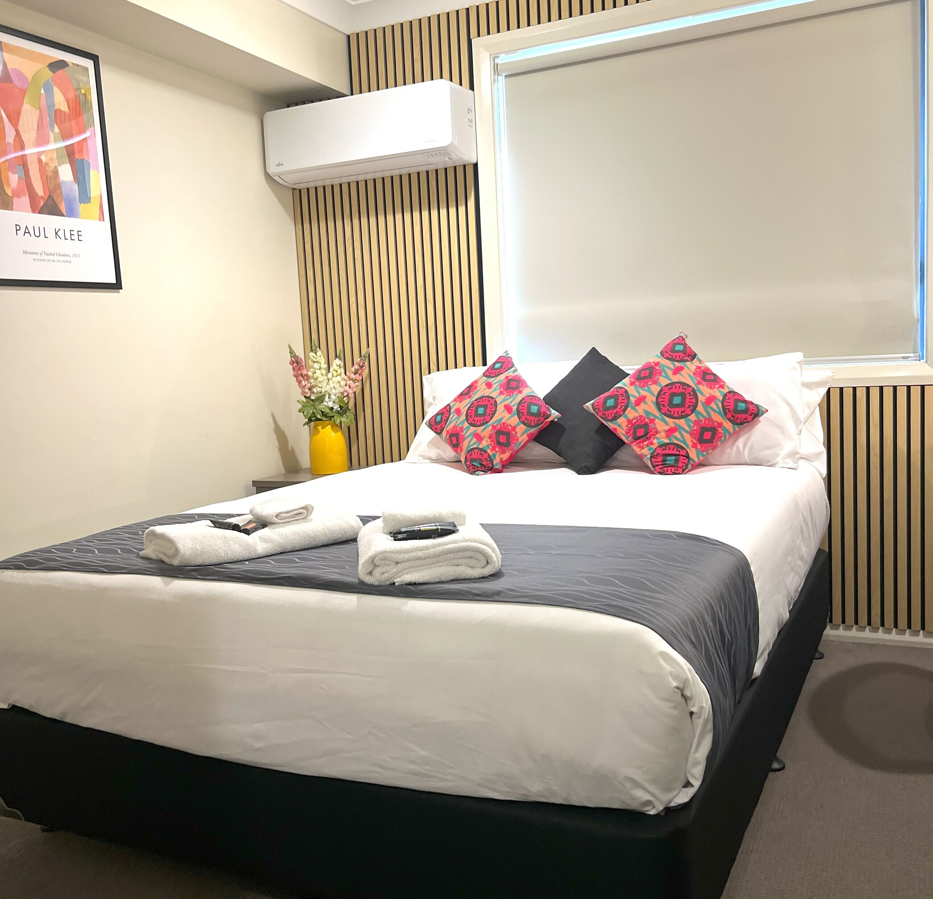 A neatly made bed with white and gray linens, two decorative pillows with a geometric pattern, a yellow vase with flowers, and a framed artwork on the wall.