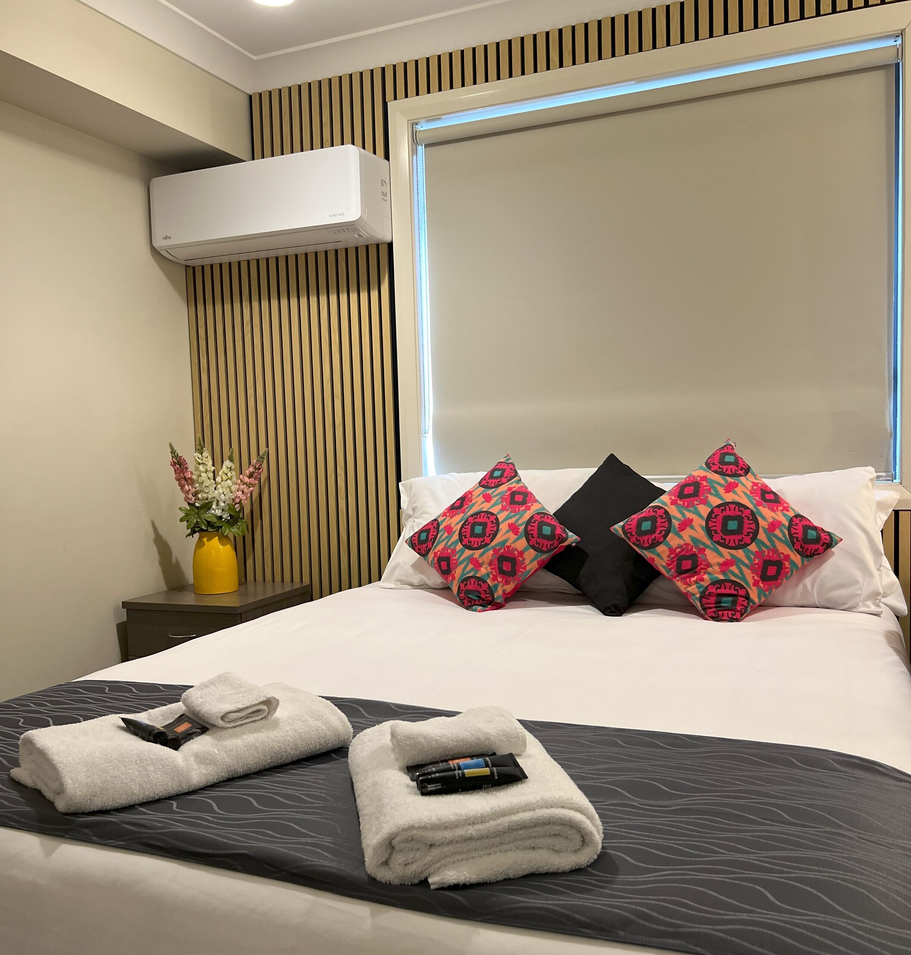 A neatly made bed with patterned pillows, a bedside table with a vase of flowers, and an air conditioning unit on the wall.