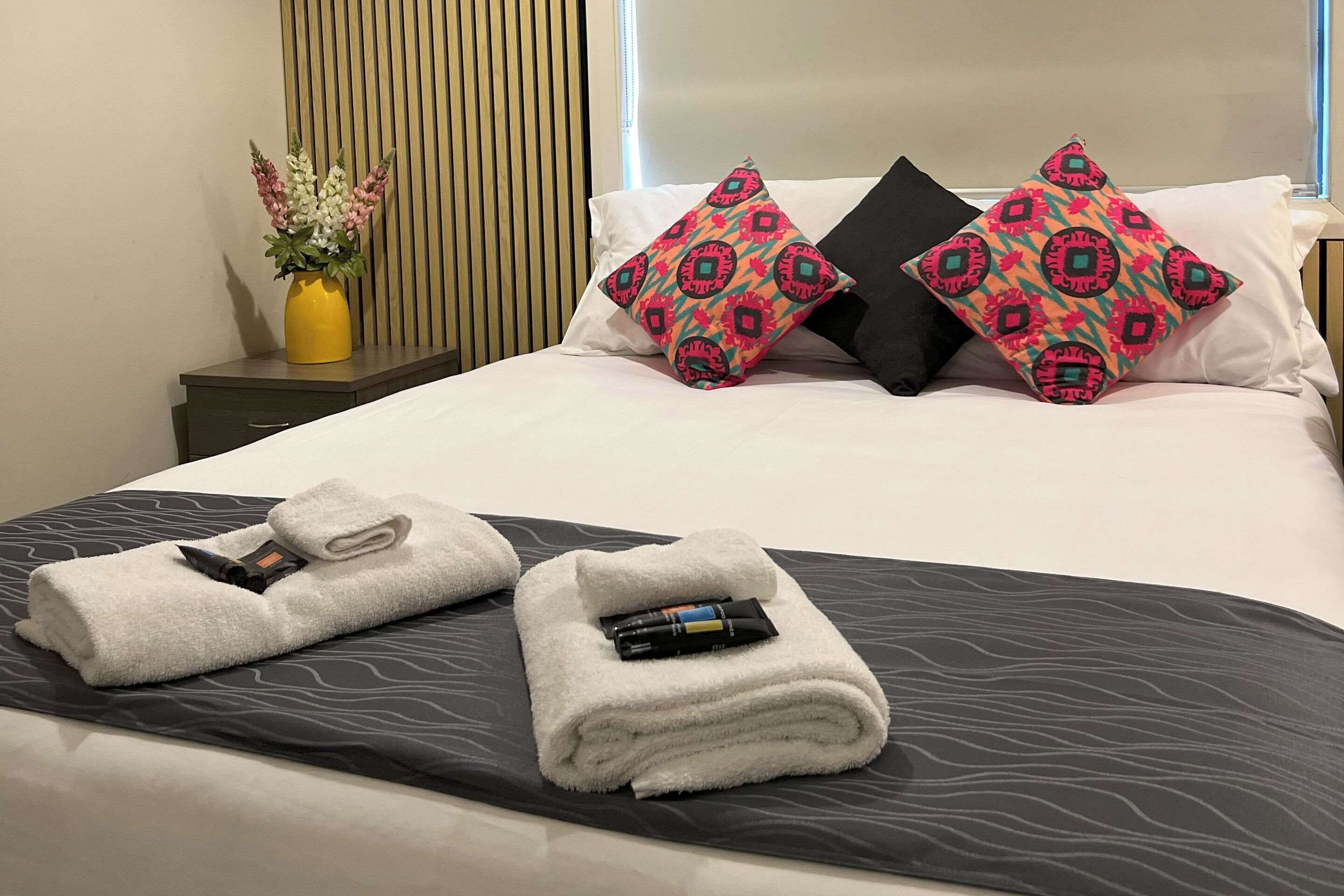 A neatly made bed with white linens, two decorative pillows, and a vase with flowers on a bedside table.
