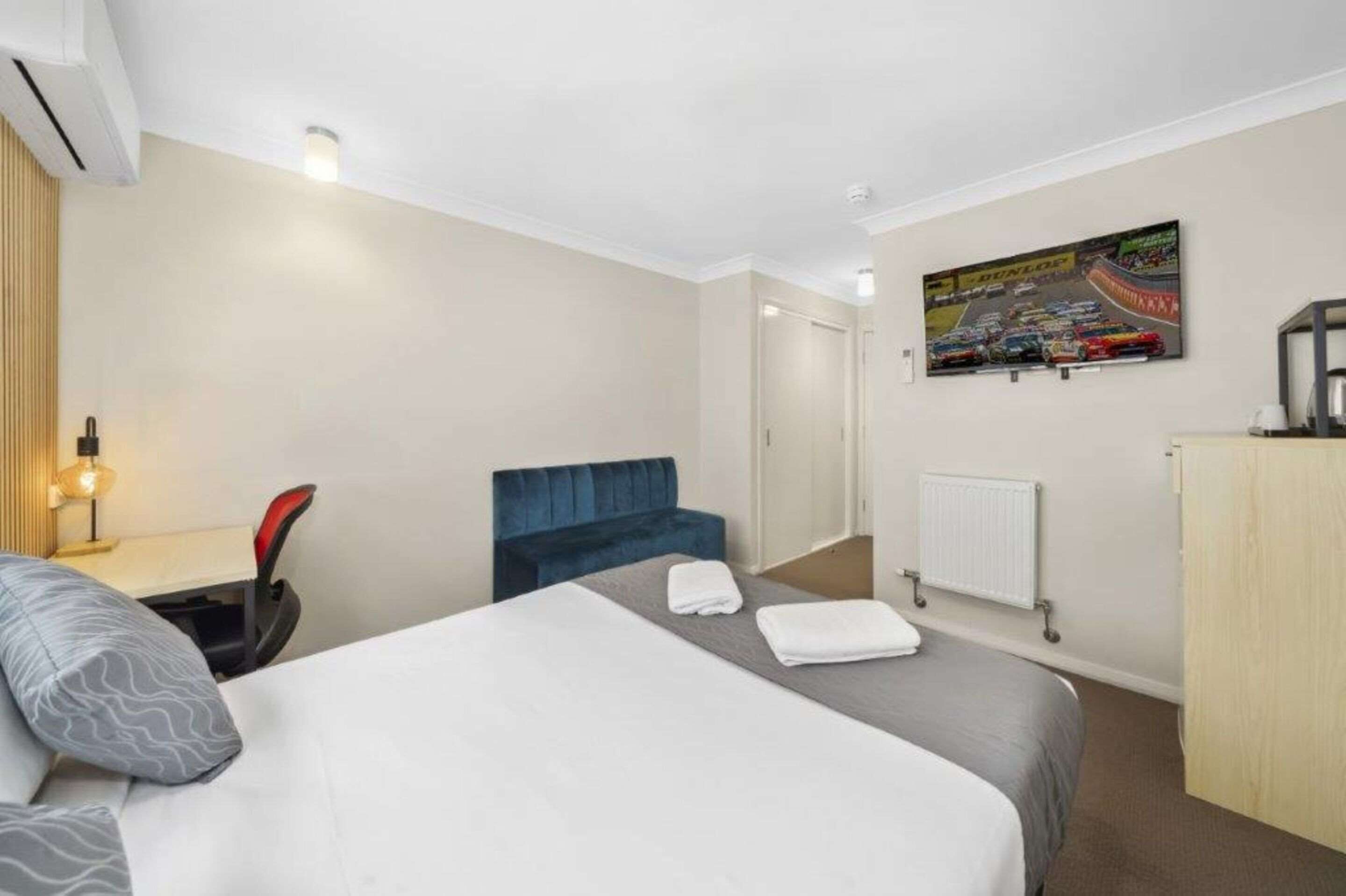 Hotelkamer met een bed, een bureau met stoel, een blauwe fauteuil, een radiator en een tv aan de muur met een racedebuut.