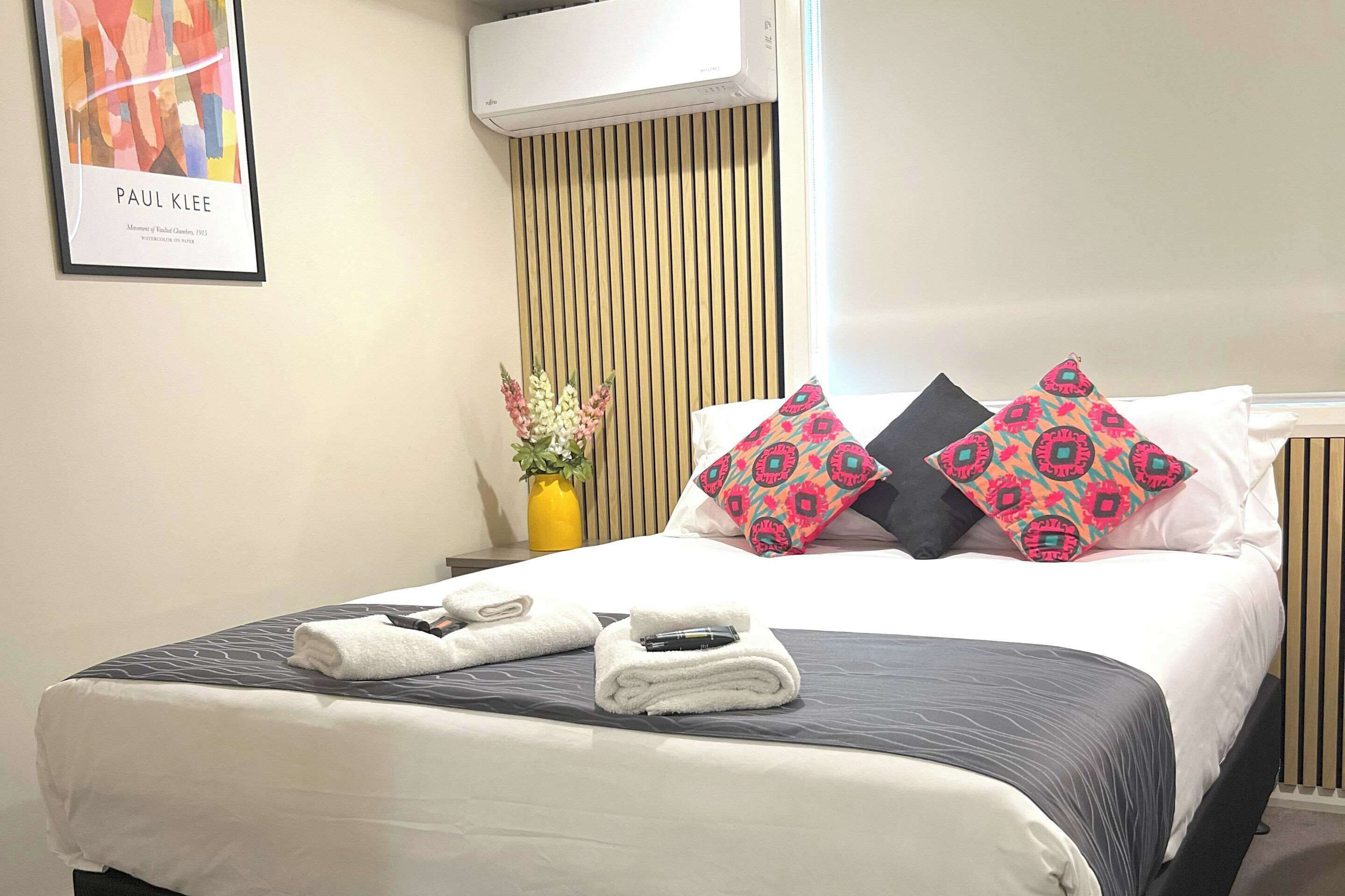 Una cama bien hecha con una colcha gris y blanca, dos almohadas decorativas y un pequeño florero con flores en la mesita de noche.