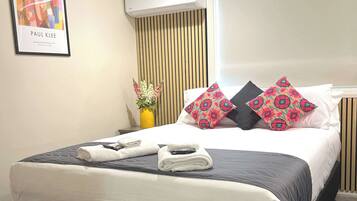 Una cama bien hecha con una colcha gris y blanca, dos almohadas decorativas y un pequeño florero con flores en la mesita de noche.