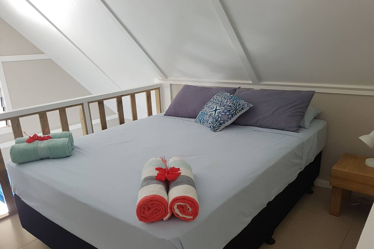 Una cama bien hecha con toallas enrolladas y una almohada decorativa, una mesita de noche de madera y vistas al exterior.