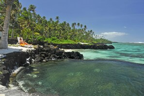 A beach with clear turquoise water, a rocky shoreline, and a person relaxing on a lounge chair. - Sa'Moana Beach Bungalows (Salamumu)
