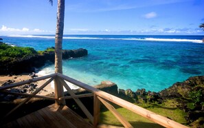 Eine Holzterrasse mit Blick auf einen klaren, blauen Ozean, eine Palme, eine felsige Küstenlinie und den Strand.