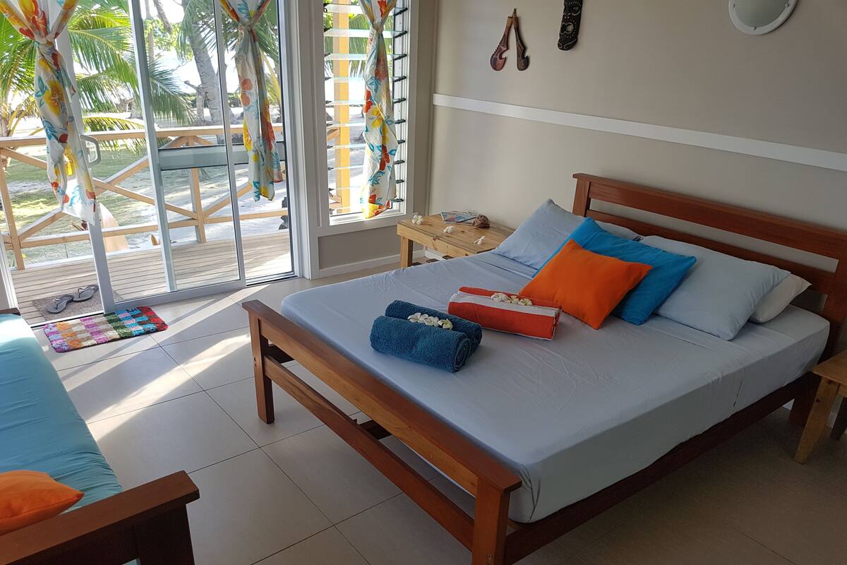 Un dormitorio con cama de madera, almohadas coloridas y vistas a una terraza de madera y palmeras.