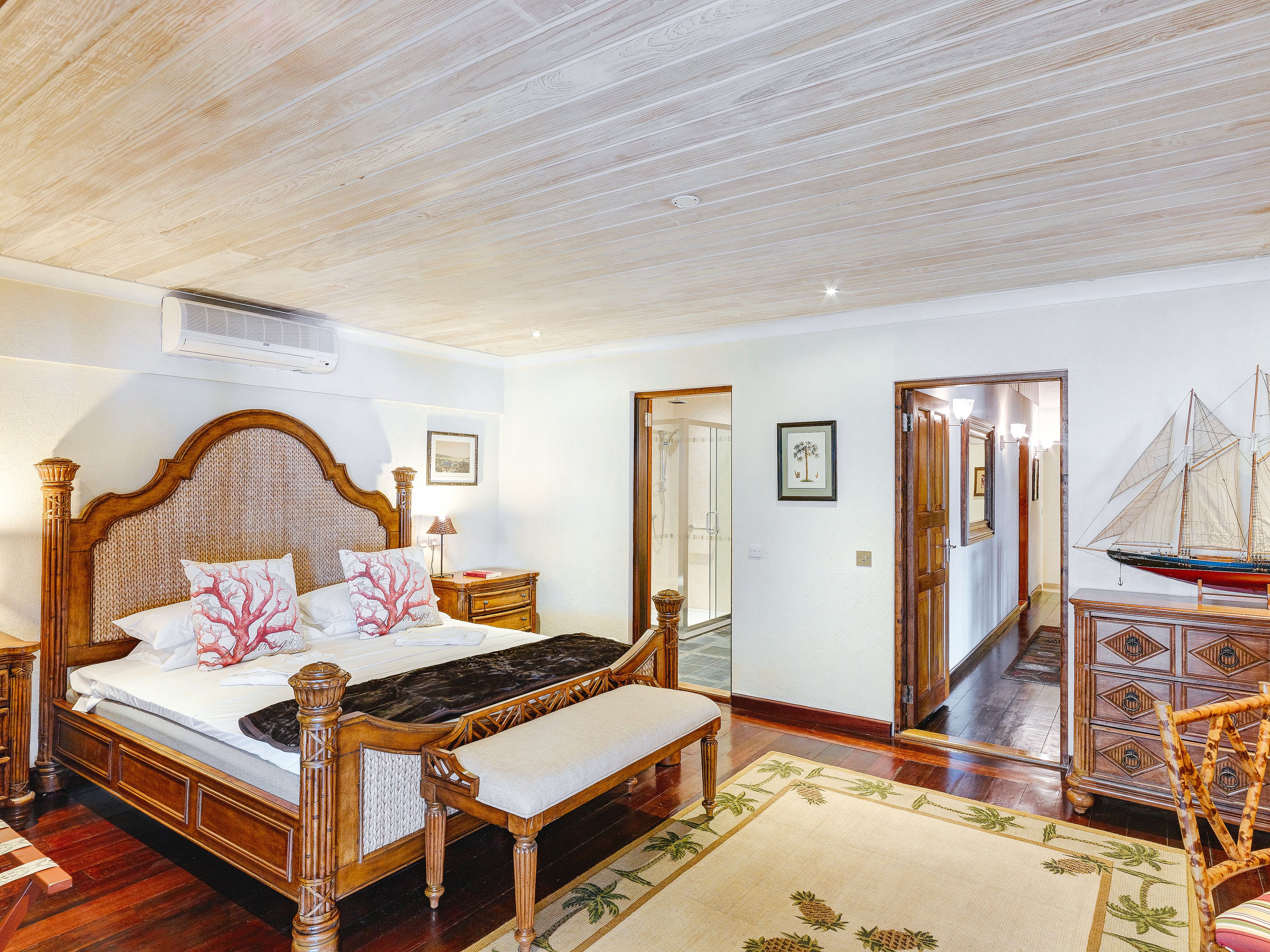 A bedroom with a wooden bed, a bench, a small table with a sailboat model, and a shower area.