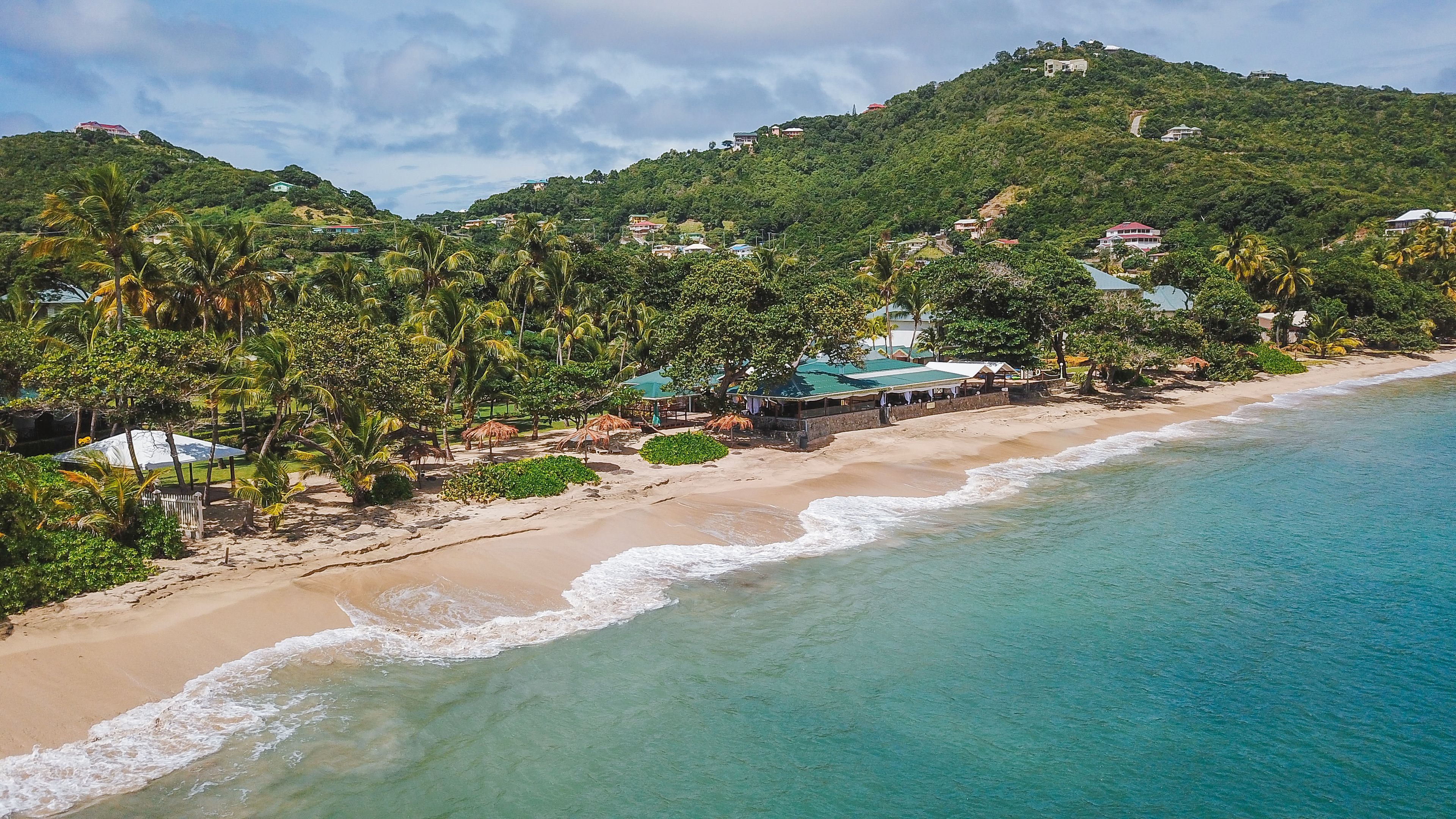 A beach with clear turquoise waters, a sandy shore, and a backdrop of lush green hills with scattered buildings.