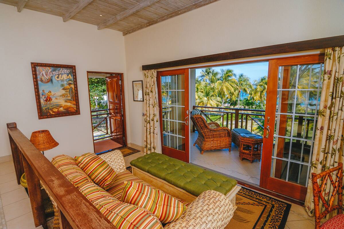 A room with a sofa, a green ottoman, wicker chairs, and a view of palm trees through large sliding glass doors.