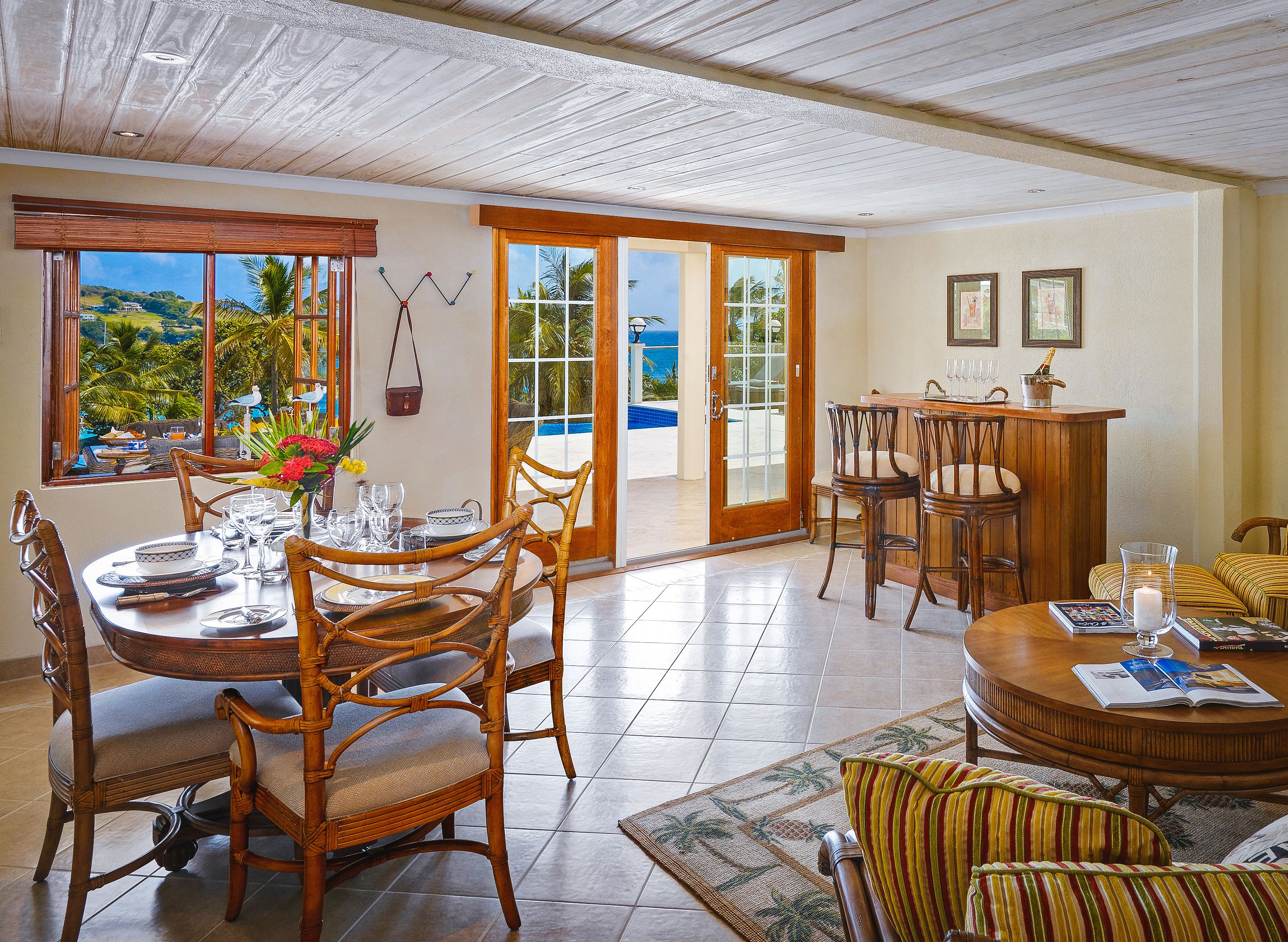 A dining area with a table set for four, surrounded by wicker chairs, a wooden bar, and a view of the ocean through large windows.