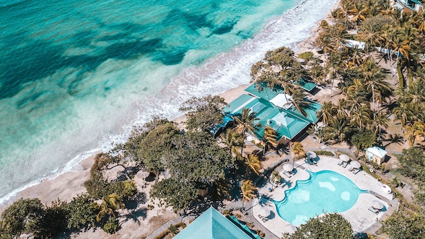 Vista aérea de un complejo turístico en la costa con aguas turquesas cristalinas, una playa de arena y una zona de piscina rodeada de palmeras.
