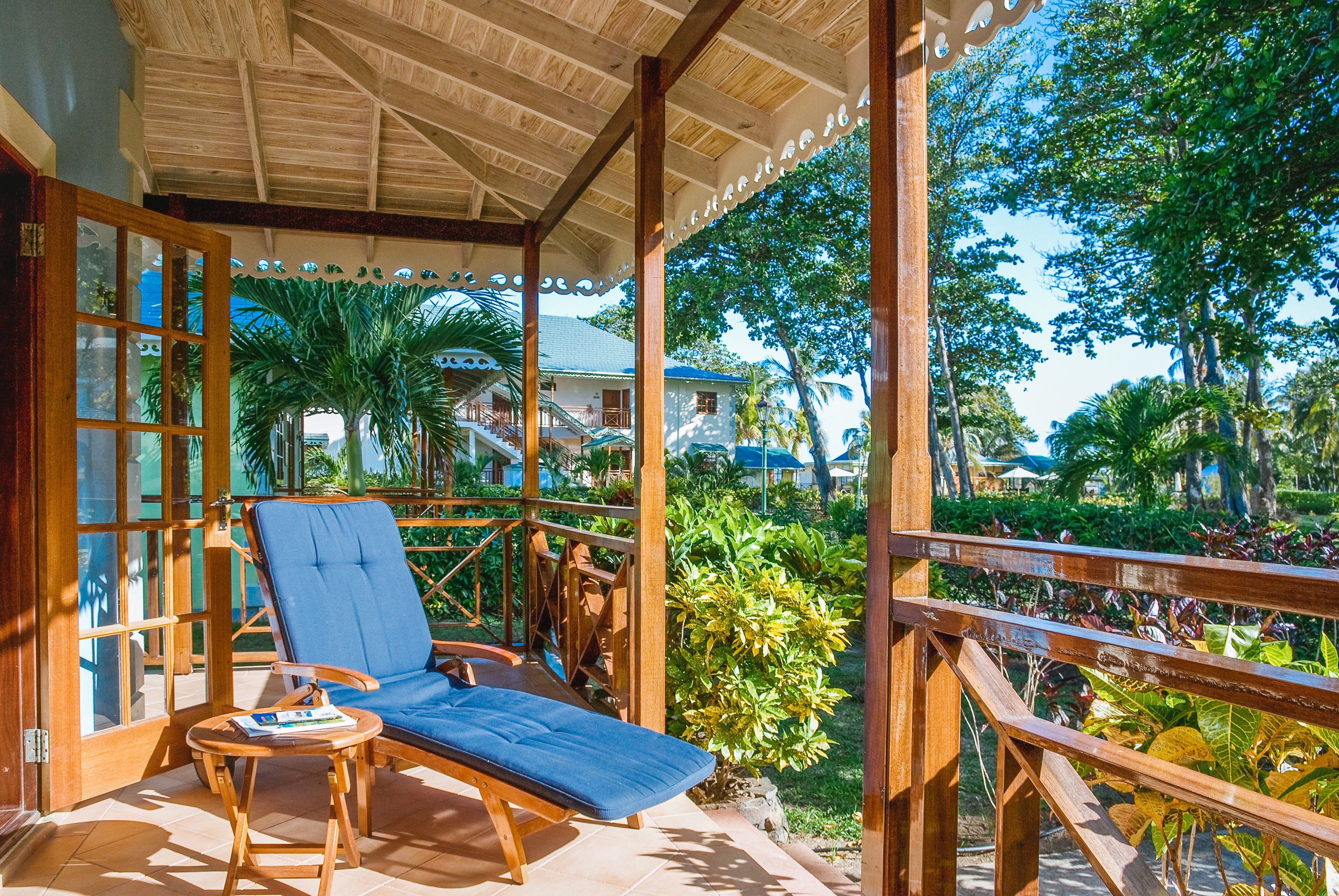 Una terraza de madera con una silla lounge azul y una mesita, con vistas a un jardín exuberante lleno de plantas y un edificio lejano.
