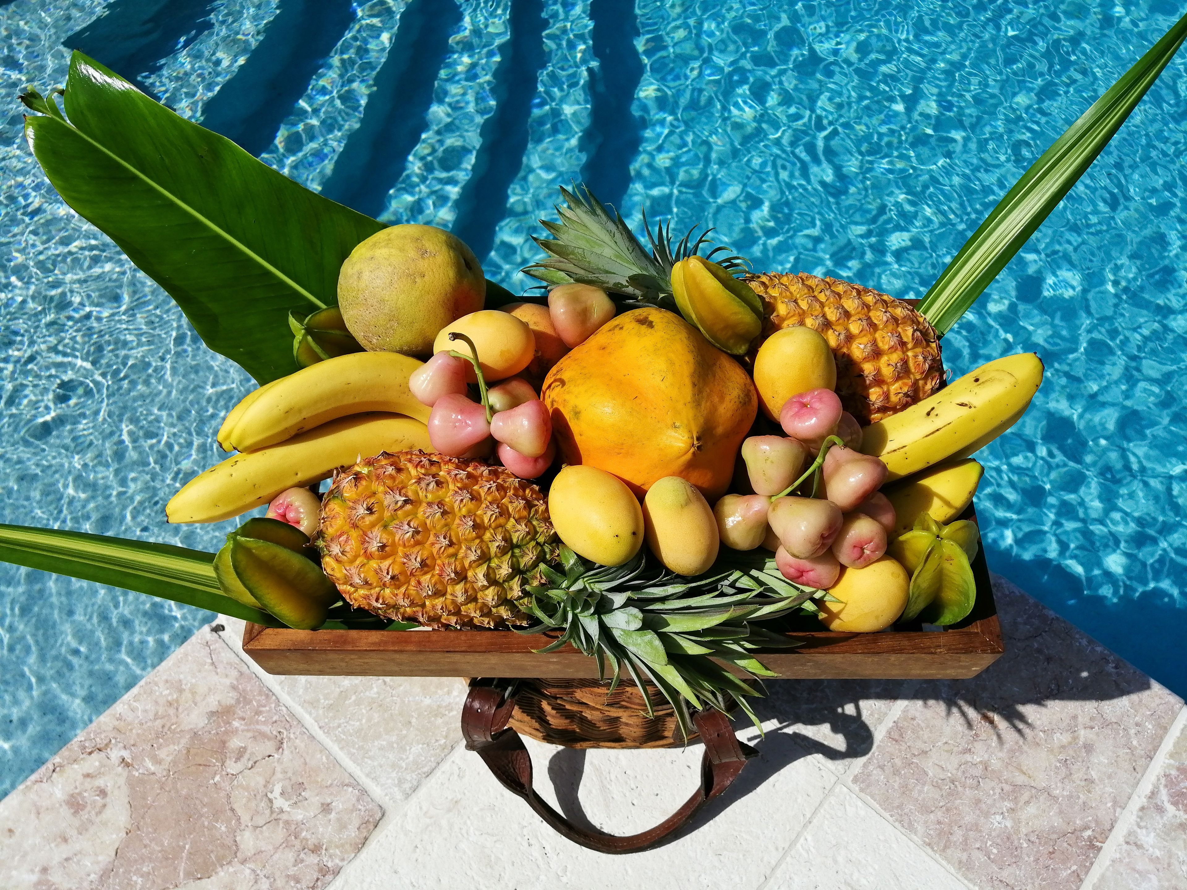 Cestino di frutta con banane, ananas, mango e papaya vicino a una piscina.