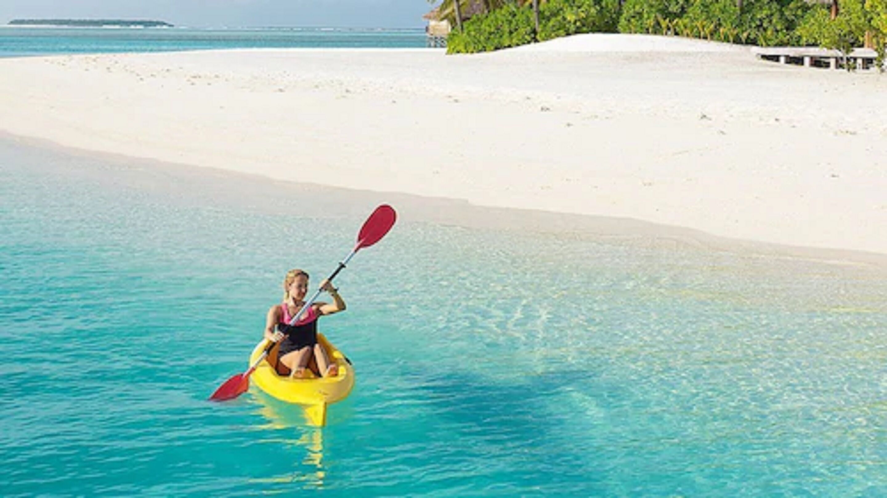 A person kayaking on clear turquoise water near a white sandy beach with a small island in the distance.