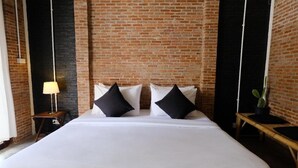 Une chambre d’hôtel avec un mur en briques, un lit avec une literie blanche et des oreillers noirs, une petite table avec une lampe et une plante en pot.