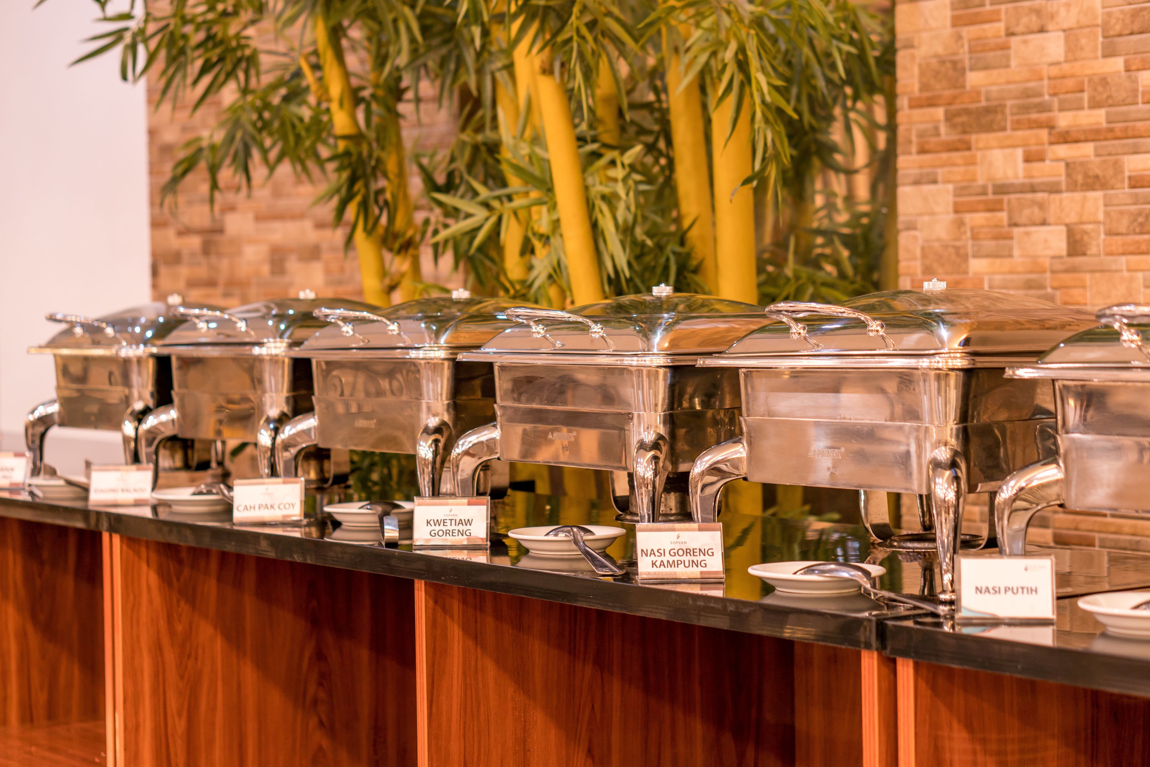 A buffet line with clear labels for various dishes, a brick wall background, and a bamboo plant.