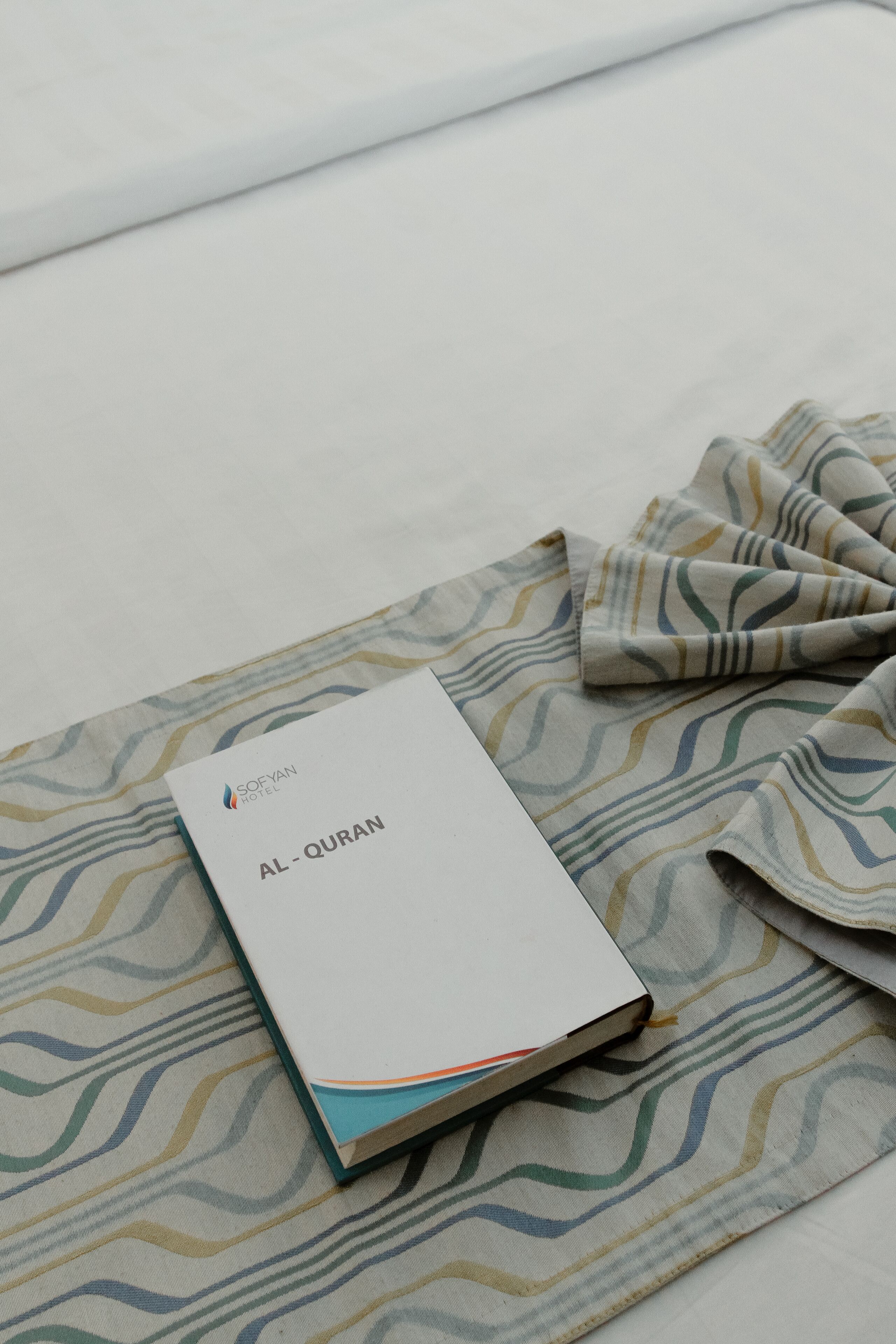 A hotel room with a bed, a patterned bedspread, and a book titled "AL-QURAN" on the bed.