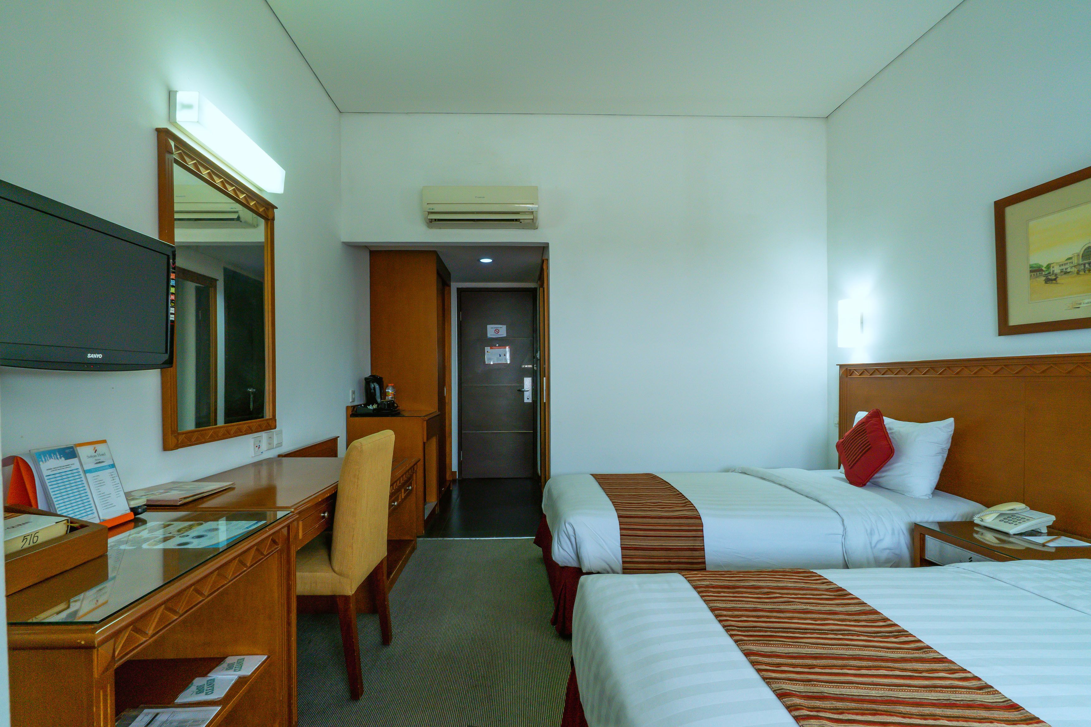A hotel room with two beds, a desk, a chair, a TV, and a door leading to a hallway.