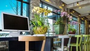 Un interior moderno con un monitor grande, una mesa de madera y sillas, rodeado de ventanas amplias y plantas en macetas.