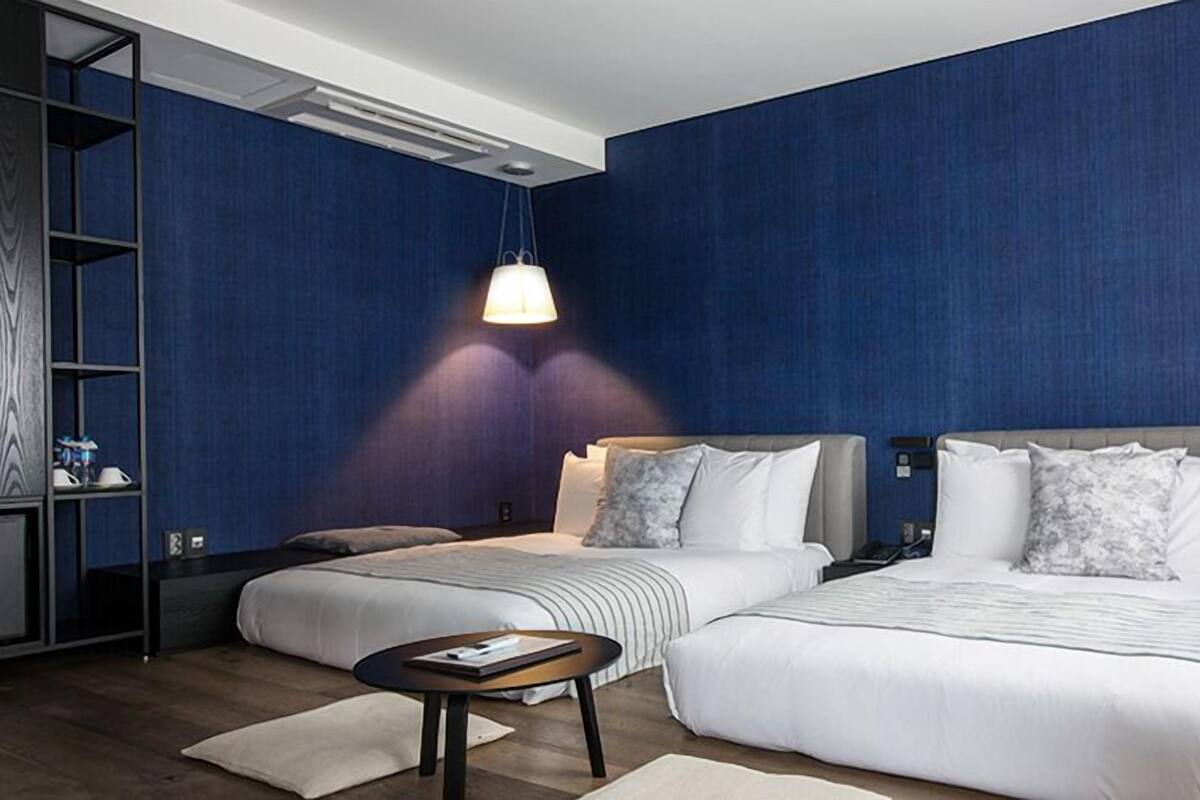 A hotel room with two beds, a nightstand, a desk, and a shelving unit.