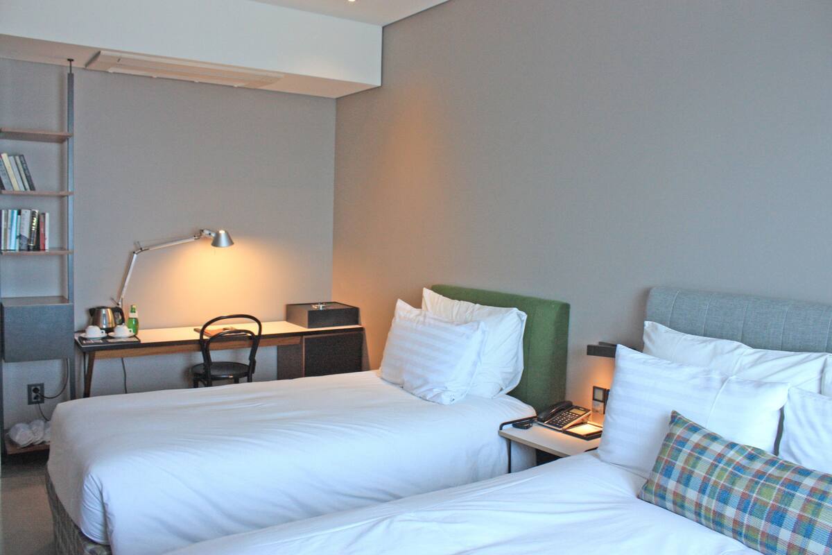 A hotel room with two beds, a desk with a lamp, a chair, a bookshelf, and a nightstand with a phone.