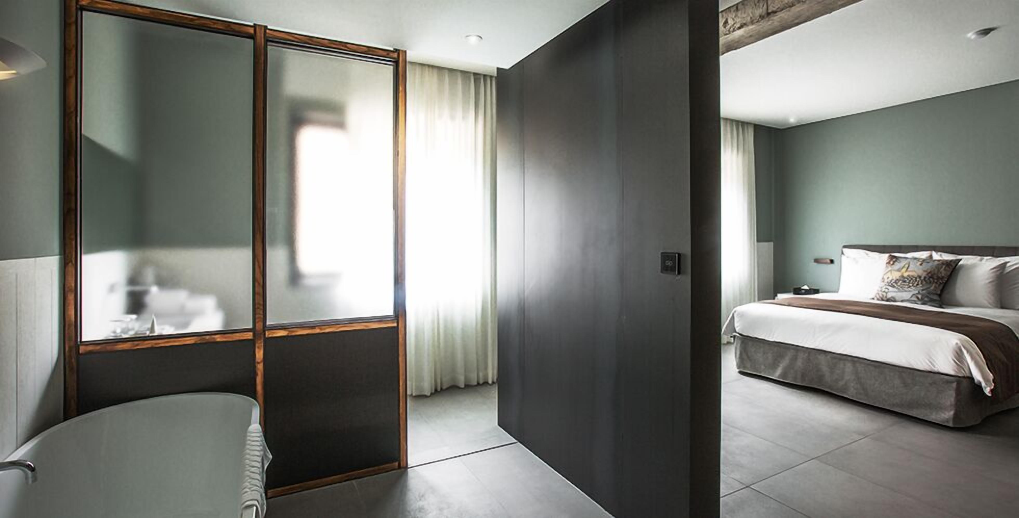 A modern hotel room with a large bed, a separate bathroom with a bathtub, and a sliding door leading to another room.