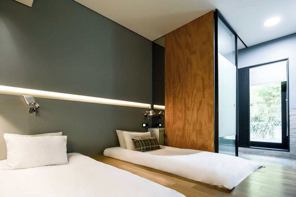 A modern bedroom with two beds, a wooden accent wall, and a large window offering a view of greenery.
