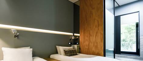 A modern bedroom with two beds, a wooden accent wall, and a large window offering a view of greenery.
