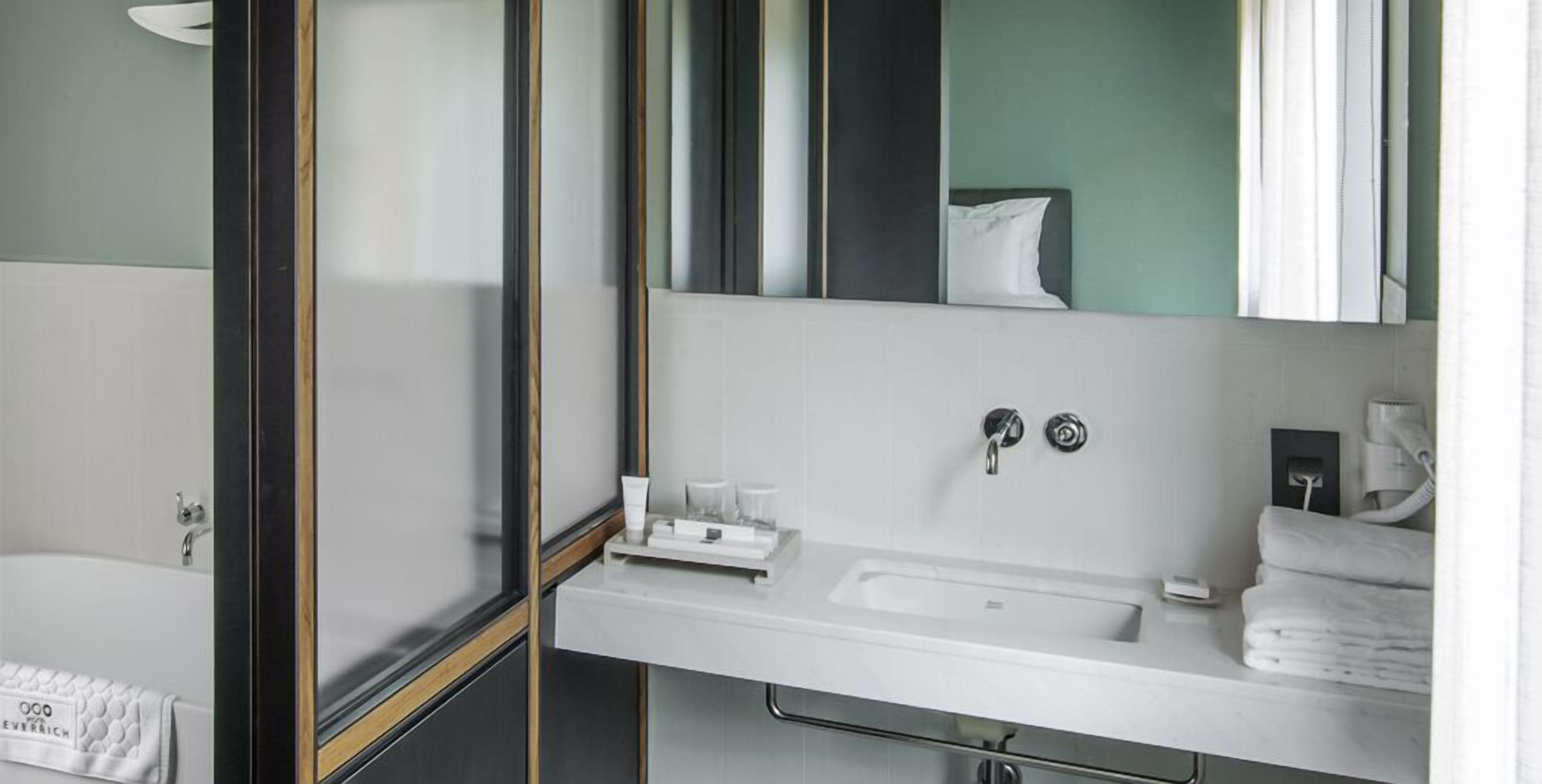 A modern bathroom with a white sink, two faucets, and a mirror. There is a towel rack with folded towels and a small tray with toiletries.