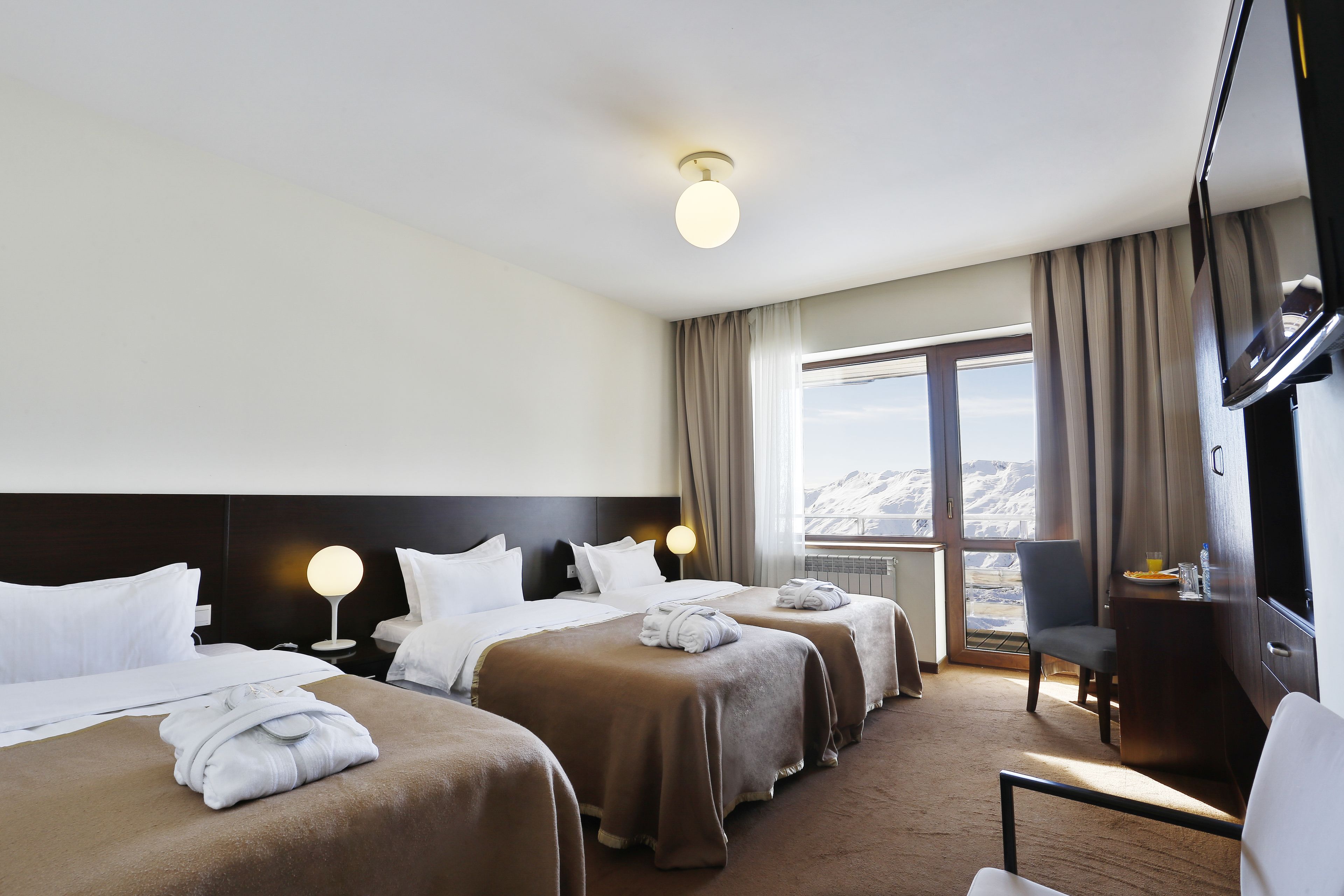 A hotel room with two beds, a desk, a chair, a TV, and a large window with a mountain view.