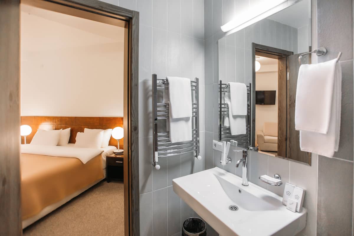 Habitación de hotel con cama, lámparas de noche, baño con lavamanos, espejo, toallero y un cubo de basura.