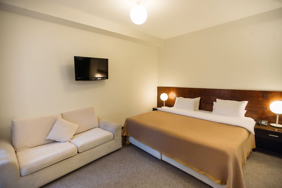 Habitación de hotel con cama, sofá, televisor montado en la pared y mesita de noche con lámpara.