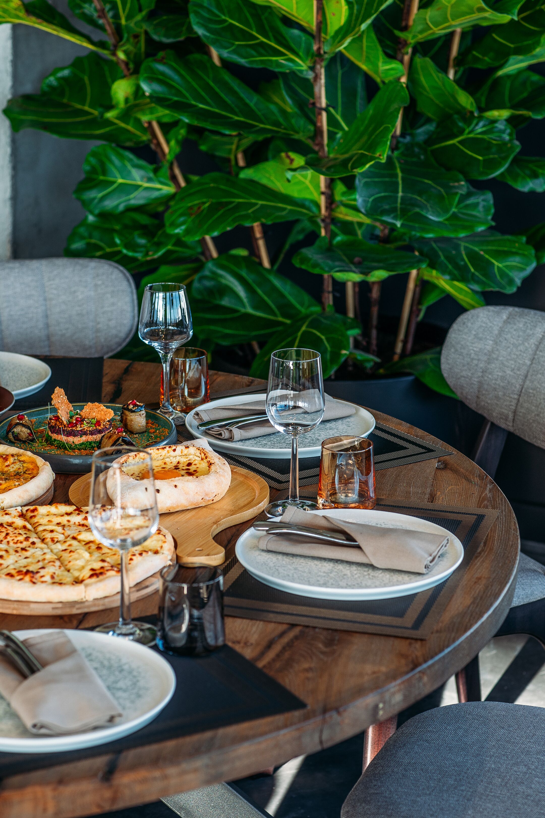 A round dining table set with plates, glasses, and cutlery, surrounded by chairs and a potted plant.