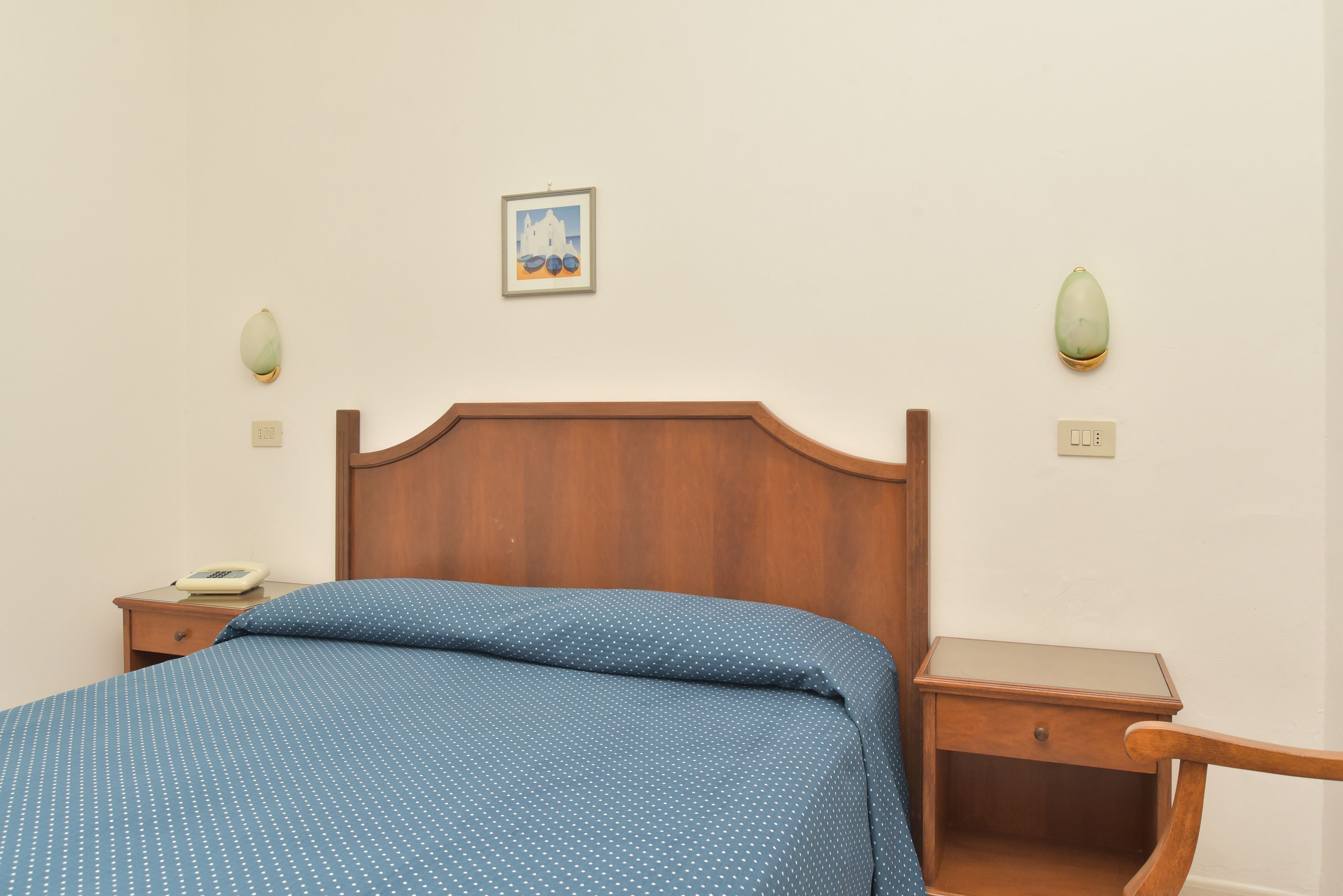 A bedroom with a wooden headboard, a blue polka dot bedspread, a bedside table with a phone, and a small picture on the wall.
