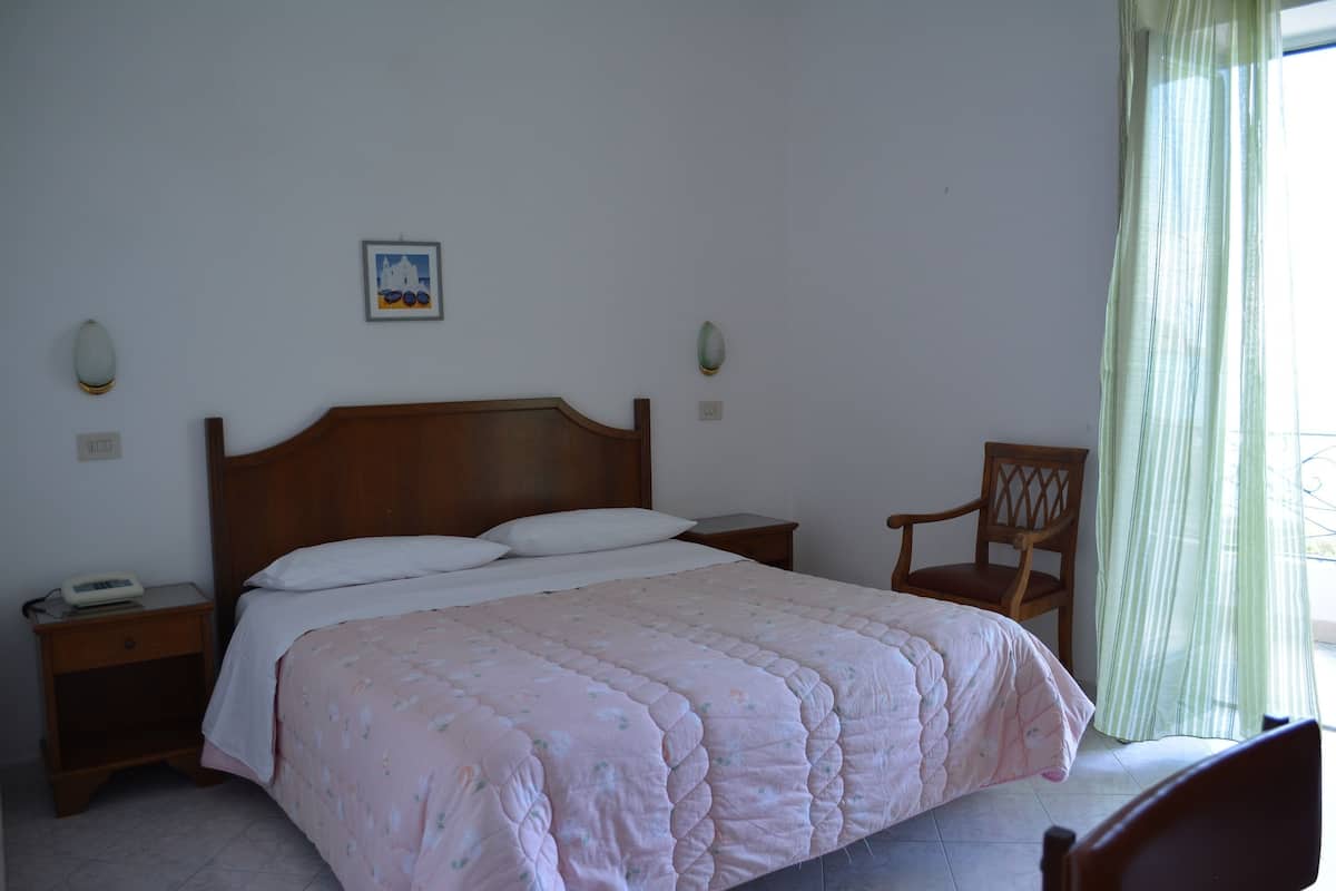 A bedroom with a wooden bed, a bedside table with a lamp, a chair, and a small picture on the wall.