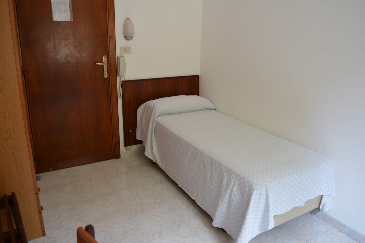 A small, single-bed room with a wooden door and a bedside telephone.