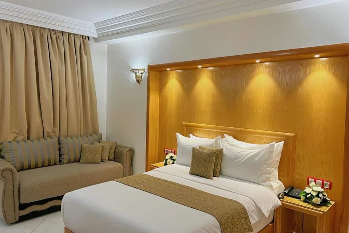 A hotel room with a large bed, a sofa, a nightstand with a phone, and a wall-mounted light fixture.