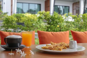 Una mesa con una taza de café, un vaso de jugo de naranja y un plato de comida, frente a un fondo de vegetación y cojines anaranjados.