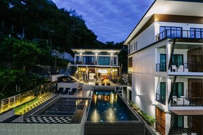 Un edificio moderno de varios pisos con piscina, zona de estar exterior y una entrada bien iluminada.