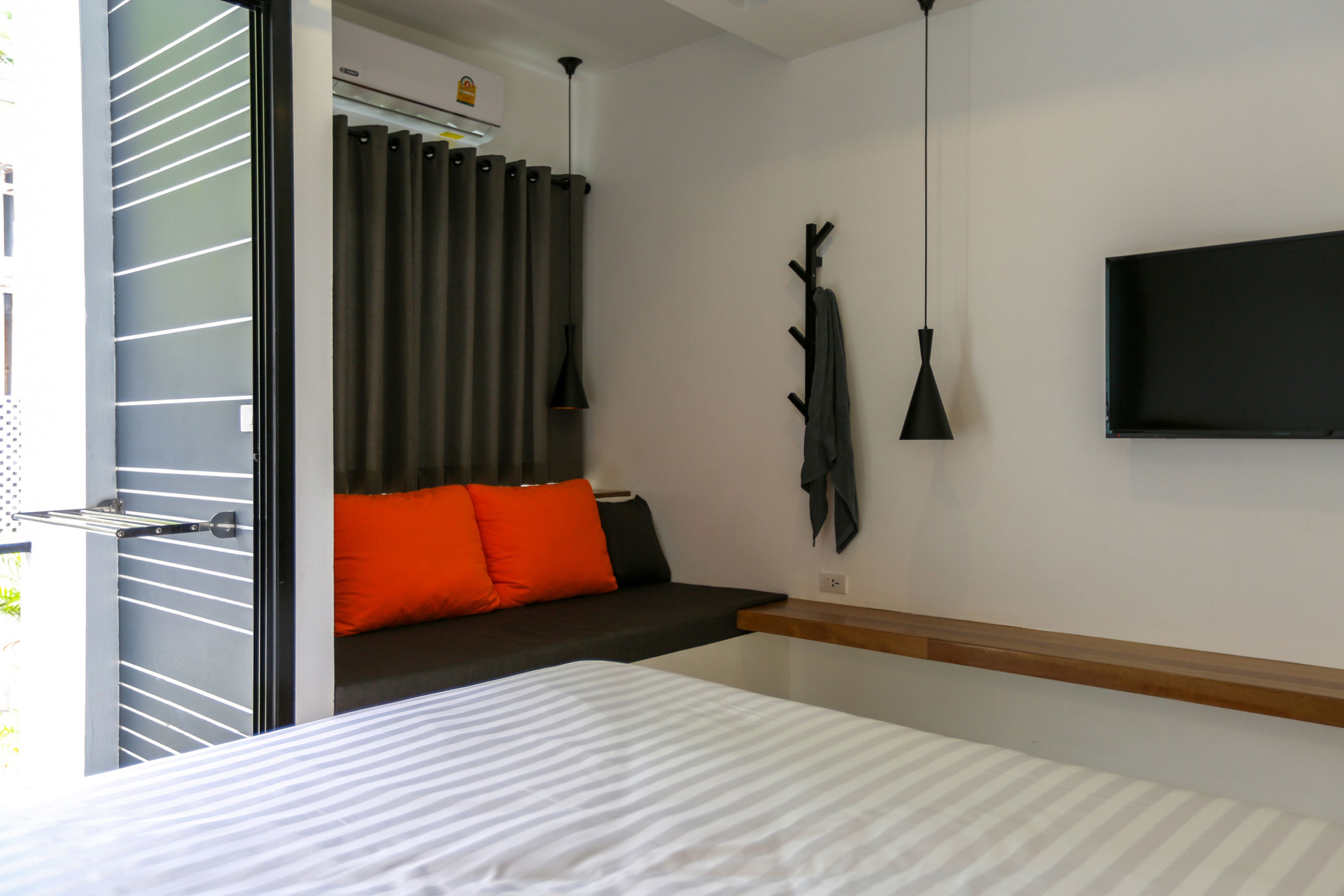 Una habitación de hotel moderna con una cama, una zona de estar con cojines naranjas, un televisor en la pared y un perchero.