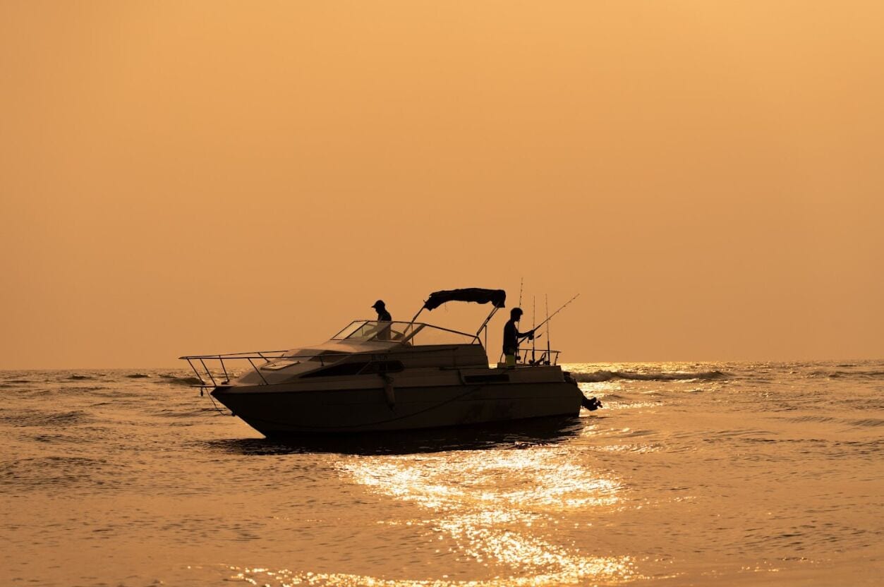 A boat with two people on deck, sailing on calm waters during sunset.