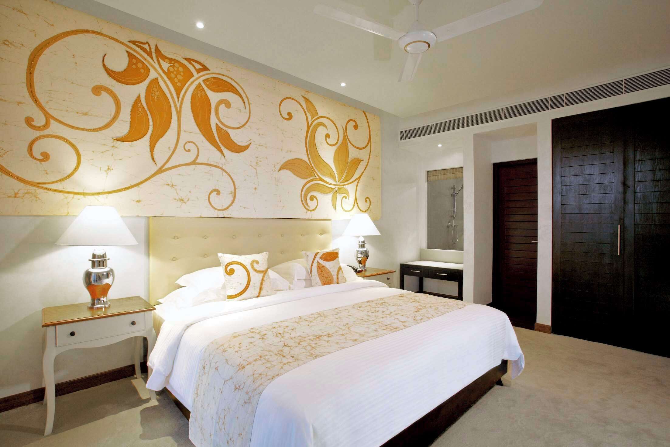 A hotel room with a large bed, two bedside tables with lamps, a decorative wall mural, a dark wooden wardrobe, and a ceiling fan.