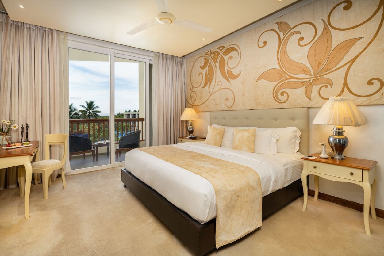 A spacious bedroom with a large bed, a desk, and a chair, a balcony with a view, and a decorative wall.