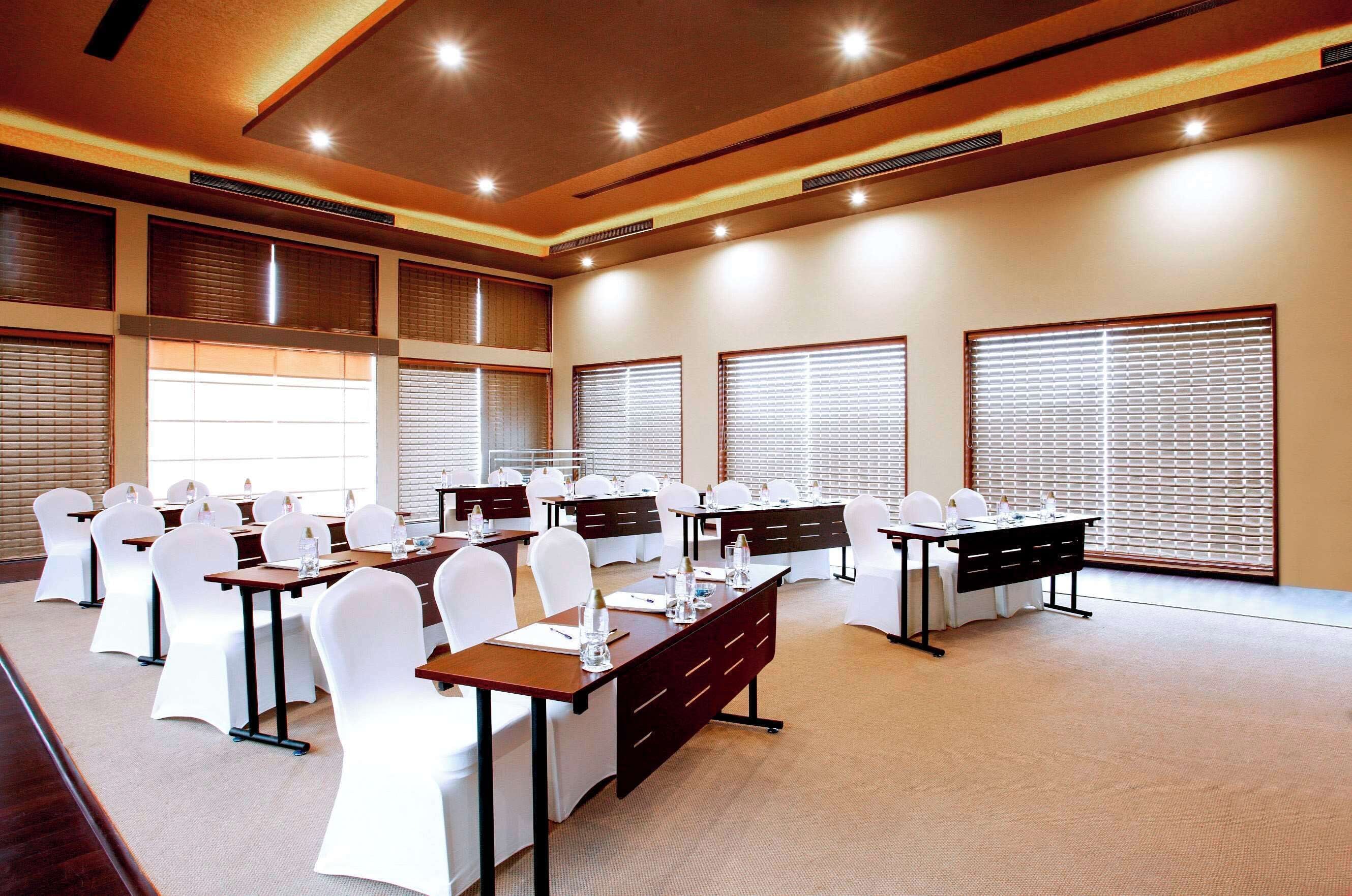 A conference room with multiple tables, white chairs, and large windows with blinds.