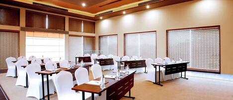 A conference room with multiple tables, white chairs, and large windows with blinds.