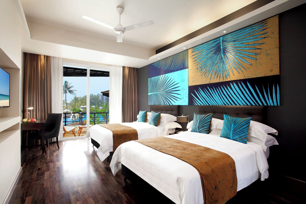 A hotel room with two beds, a desk, a chair, and a view of the beach.
